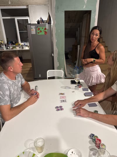In Verkhniy Yurt, Russia, four individuals are gathered around a white table, engaged in a game of poker. The room appears to be undergoing renovation, with exposed walls and construction materials visible in the background. The table is set with playing cards, poker chips, and a glass of white wine.
The individuals are:
* A man on the left, wearing a grey t-shirt with a floral pattern, is holding cards and looking intently at the game.
* A woman in the center, dressed in a black crop top and a light pink skirt, is looking towards the camera with an expressive gaze.
* Two other individuals are partially visible, their hands on the table, suggesting active participation in the game.
The scene captures a moment of casual social interaction amidst an unfinished setting, highlighting a blend of domesticity and ongoing construction.