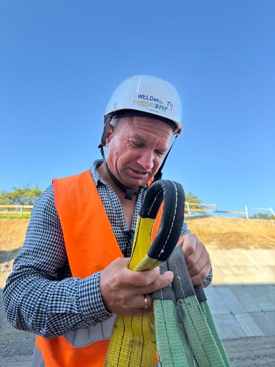 A man wearing a white hard hat with blue writing, an orange safety vest over a checkered shirt, and a grey face mask is holding a yellow and green lifting strap. He is looking down at the strap with a pained expression. The background shows a clear blue sky, a dirt embankment, and some trees. The scene appears to be outdoors, possibly at a construction site. The image was taken in Stróża, Poland.