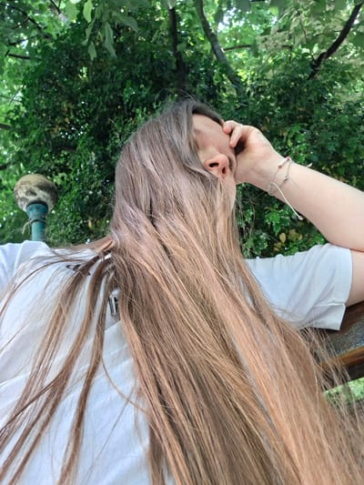 A woman with long, light brown hair is sitting outside, possibly in Galabovo, Bulgaria. Her hair is partially covering her face, and she is holding her head with her hand. She is wearing a white t-shirt and two bracelets. Behind her are green trees and foliage, which is the environment. There is also some kind of lamp nearby.