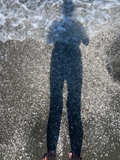 The image shows a shadow of a person standing on a beach with dark sand mixed with small shells. The person's feet are visible at the bottom of the frame, with painted toenails. Waves with white foam are lapping at the shore. The location is identified as Shekvetili, Georgia.