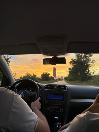 The image is a POV shot from the interior of a car, taken from the passenger seat. The car is driving down a road at sunset, with the sky filled with vibrant orange and yellow hues. A person is seen driving the car, with their hands on the steering wheel. The dashboard displays the time as 20:02 and the speed as 91. A car air freshener hangs from the rearview mirror. The car is surrounded by trees and vegetation, suggesting a rural or semi-rural setting. In the distance, there appear to be some industrial structures. The location is identified as Balanovo, Bulgaria.