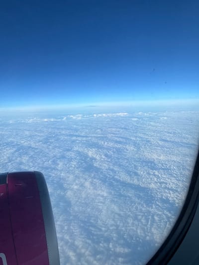 An aerial view from an aircraft window shows a dense, expansive layer of white clouds extending to a distant horizon under a clear blue sky. In the lower-left quadrant, a portion of an aircraft engine nacelle is visible; it is predominantly magenta/purple with a black leading edge and partial text "EP" on its surface. The cloud layer below appears textured, exhibiting variations in light and shadow, suggesting a stratocumulus or altocumulus formation. The sky transitions from a lighter blue at the cloud-sky interface to a deeper blue overhead. No ground features of Caselle Torinese, Italy, are discernible due to the complete cloud cover; the image depicts an atmospheric scene consistent with an aircraft operating in the vicinity of Caselle Torinese. The scene is illuminated by bright, clear daylight.