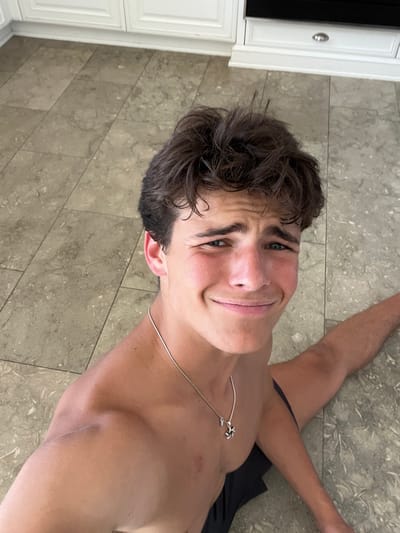 A young adult male, appearing in his late teens or early twenties, is depicted shirtless, performing a full leg split on a tiled floor. He has short, messy, dark brown hair and blue eyes, with a grimacing or forced smiling expression directed towards the camera. He wears a thin silver chain necklace with a small, stylized silver pendant. Dark fabric is visible around his waist, consistent with shorts. The flooring consists of large, light-colored square tiles with subtle marbling and visible grout lines. The background reveals a modern kitchen area with white shaker-style cabinets, silver handles, and a dark-colored built-in appliance with a silver handle above a lower cabinet. The scene is brightly lit by ambient light. This activity is observed within a private residence in the city of Rancho Palos Verdes, United States.