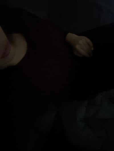 The image presents a dimly lit indoor scene featuring a single individual partially visible. The subject appears to be reclining or lying down, oriented diagonally within the frame. On the far left, the lower portion of a light-skinned face is discernible, specifically the chin and mouth with pink, slightly parted lips. The individual wears a dark, long-sleeved garment. Towards the middle-right of the frame, a light-skinned hand, appearing partially clenched, rests on an indistinct dark surface. In the lower right quadrant, a crumpled dark bluish-gray fabric, possibly a blanket or sheet, is faintly visible. The overall environment is characterized by very low light conditions, rendering most background details indistinct. There are no discernible features in the image to confirm a specific location within the city of Albi, France.
