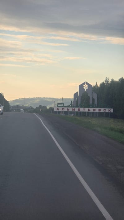 The image displays an asphalt road extending into the distance under a sky with a low sun angle, suggesting either early morning or late afternoon. The scene is located near Nizhnyaya Maktama, Russia, as specified, though a prominent sign indicates the city of Almetyevsk.
In the foreground, a dark grey asphalt road occupies the lower portion of the frame, showing signs of dampness. A solid white line delineates the right edge of the road, with no central lane markings visible. On the right side of the road, a grassy verge supports a large welcome sign. This sign features a grey, multi-part architectural structure with geometric forms, centered by a stylized emblem. Below this structure, the name "АЛЬМЕТЬЕВСК" (Almetyevsk) is spelled in red Cyrillic letters against a white background. Beyond the sign, a dense line of dark green trees forms a forested edge.
On the left side of the road, a portion of a white vehicle, possibly an SUV or large car, is visible in the near distance, traveling in the same direction as the viewer. Further along the left side, a series of horizontal pipe-like structures are visible above the terrain. Rolling hills, covered with lighter green vegetation and appearing hazy, form the distant background on the left. The sky overhead transitions from a lighter blue at the horizon to darker grey tones higher up, with several elongated, stratified clouds ranging in color from pale cream to light grey. No individuals are visibly engaged in direct actions or interactions within the frame.
