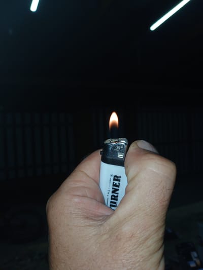 A close-up shot captures a hand holding a white lighter with black lettering, from which a small flame emerges. The thumb of the hand is positioned to operate the lighter's ignition mechanism. The lettering on the lighter is partially visible, reading "THE TURNER". The background is dark and out of focus, with horizontal lines suggesting blinds or a similar slatted surface. Two streaks of light are visible in the upper left portion of the frame, possibly from a flash or other light source. The scene appears to be indoors, given the presence of the blinds, and the overall lighting suggests it might be nighttime or a dimly lit environment. The image focuses on the action of igniting the lighter.