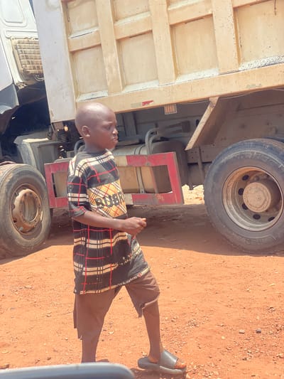 A person wearing brown shorts and a plaid shirt stands next to a large truck on a dirt surface in Kwali, Nigeria. The person wears black sandals on their feet. The truck has two visible tires, and a beige rectangular component is visible on the left side of the image. The surface is reddish-brown dirt with some small rocks or pebbles.