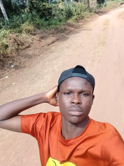A young man with dark skin and a black baseball cap faces the camera. He wears an orange t-shirt with a partially visible yellow design. His left hand rests on his head near his ear. The background features a dirt road surrounded by green vegetation. The sunlight is bright, suggesting a sunny day. The man appears to be outdoors.