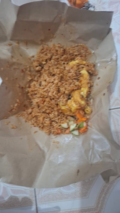 The image shows a serving of fried rice, likely for takeaway, laid out on brown paper. The fried rice is a mound of brown-tinted grains, with scattered pieces of scrambled egg mixed in. On the right side of the rice, there are small, diced pieces of vegetables, appearing to be cucumber and carrot. A small key chain with an orange charm is visible on the left edge of the image. The food is placed on a surface that appears to be tiled, with a pattern of light pink and cream colors. The lighting suggests it's daytime, possibly indoors due to the indoor setting indicated by the tile pattern and the lack of natural outdoor light. The overall presentation is simple and casual, typical of street food or a quick meal. The food appears to be the main subject, with no people visible. The scene is confined to the food and its immediate surroundings. There is no visible text in the image. The context places this scene in Batam, Indonesia.