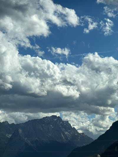 The image depicts a dramatic mountain landscape under a dynamic sky. A range of jagged, snow-capped mountains dominates the lower half of the frame, their dark, rocky slopes contrasted by patches of white snow. In the foreground, several horizontal wires stretch across the scene, suggesting a power line or fence. Above the mountains, a vast expanse of blue sky is filled with a complex arrangement of clouds. Large, fluffy cumulus clouds, some brightly lit and others casting deep shadows, create a sense of depth and texture. The overall impression is one of natural grandeur and a powerful, ever-changing atmosphere. This scene is consistent with views from Farchant, Germany, a Bavarian municipality known for its proximity to the Alps and its stunning mountain scenery.