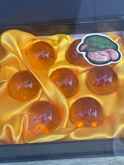 A black box with a dark blue lid is open, revealing a yellow satin lining. Six orange, transparent spheres are nestled within the satin. Each sphere appears to have red stars inside, with the number of stars varying. A sticker of a pig wearing a green military-style hat with a red star is affixed to the right side of the box, near one of the spheres. The background is out of focus, but appears to be a surface or wall. The image is well-lit, with reflections visible on the spheres. The exact location in Getafe, Spain, cannot be determined from the provided image.