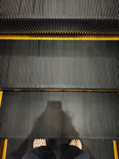 The image shows a close-up, eye-level view of an escalator's treads. The escalator is made of dark, ribbed metal with a bright yellow stripe running horizontally across it. A person's feet, clad in light-colored clogs and dark pants, are visible at the bottom of the frame, standing on the escalator. A shadow of the person is cast on the treads above. The scene appears to be indoors, with the lighting suggesting an artificial environment, common in shopping malls or large public buildings. The image composition emphasizes the repetitive pattern of the escalator treads. There are no discernible landmarks or contextual clues to place this image specifically in Pekalongan, Indonesia.