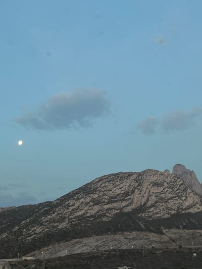The image shows a large, rocky mountain range, possibly the Sierra Madre Oriental, taken in the afternoon from a location in Ciudad Santa Catarina, Mexico, at the coordinates (25.657, -100.449). The sky is a light blue with a few white clouds and the moon, which is a bright white disc. The mountain is a light brown color with a few patches of green vegetation. There is a small amount of vegetation in the foreground, as well as a few light sources that appear to be street lights. The image is taken from a distance and the only subject is the mountain. The image is not close-up and there are no people present.