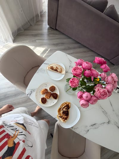 In Malinovka, Russia, a top-down view captures a domestic scene on a marble-patterned table. A bouquet of vibrant pink roses in a clear glass vase dominates the center, its blooms filling the frame with color. To the left of the flowers, a white plate holds a folded, browned flatbread. Below that, another white plate displays five small, round pancakes sprinkled with dark specks. To the right of the roses, a third white plate contains a single folded piece of browned flatbread, appearing to be the same as the one on the left. The table is set on a grey wooden floor, with the legs of a beige upholstered chair visible on the left. In the lower left corner, a person's legs are visible, clad in white shorts with a graphic print featuring a figure and stripes, partially obscuring the floor. The background reveals a section of a grey sofa and a sheer white curtain hanging near a wall-mounted outlet.