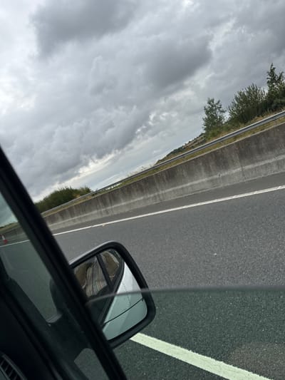 The image shows a view from inside a vehicle traveling on a highway, likely in Forristalstown, Ireland, given the context. The sky is overcast with heavy, dark clouds. The highway has a concrete barrier separating it from the opposing lanes. Vegetation, including trees and bushes, grows on the embankment behind the barrier. The road surface is asphalt, with white lane markings visible. A side mirror is positioned in the foreground, reflecting the interior of the car, which appears to contain a person wearing a striped shirt. There are traffic cones visible on the left side of the highway. The overall impression is one of transit under inclement weather conditions.