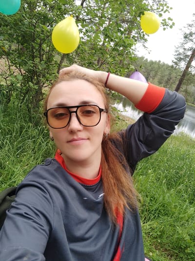 A young woman with red hair and a dark gray sweatshirt with orange trim stands outdoors in front of a body of water and trees. She is wearing large black-rimmed glasses with orange lenses. She is holding her right arm up and touching her head. Several balloons are visible in the background, including one yellow balloon hanging from a tree branch and another yellow balloon in the upper right corner of the image. A purple balloon is also visible in the background. The ground is covered in tall green grass. The location is identified as Orekhovo, Russia.