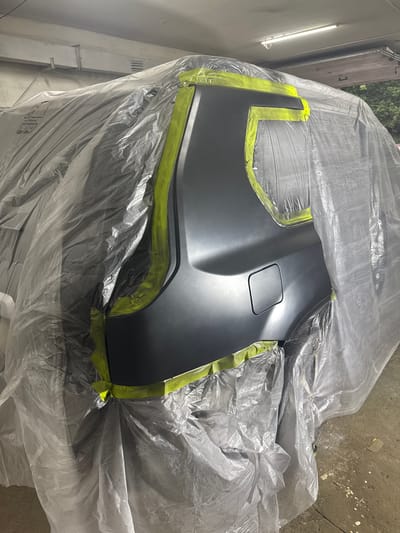 This is an overhead, eye-level shot of a dark gray car bumper that has been masked off with bright yellow painter's tape. The bumper is partially covered in clear plastic sheeting, which is draped over a larger portion of the vehicle. The plastic sheeting has a slightly dusty appearance. The bumper itself has a subtle sheen and appears to be undergoing some kind of automotive work, likely painting or repair. There is a square-shaped indentation on the surface of the bumper, possibly a fuel filler door or a similar access panel. The background is blurred but appears to be an indoor setting with concrete flooring and industrial-style lighting on the left side of the frame. The overall impression is a work-in-progress in an auto body shop or garage. There is no visible text in the image.