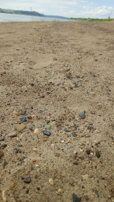 The image depicts a low-angle ground-level perspective of a sandy riverbank or beach, identified as being in the vicinity of Шолья, Russia. The foreground is in sharp focus, showcasing fine-grained, light brown sand interspersed with numerous small, multi-colored pebbles and gravel. These pebbles exhibit a range of natural colors including grey, black, white, various shades of brown, reddish-brown, green, and occasional hints of blue and orange. The sand's texture is visible, indicating individual particles.
Moving towards the midground, the sandy surface becomes progressively out of focus due to a shallow depth of field. A few faint, dark linear elements, possibly dried plant matter or debris, are minimally discernible in this area.
The background, largely out of focus, reveals a wide body of water, likely a river or lake, appearing calm with a light grey-blue hue. Beyond the water, a distant landmass characterized by gently sloping hills or banks, with patches of green vegetation and darker contours, stretches across the horizon.
The sky above the distant landmass is visible in the uppermost portion of the image, displaying a pale blue color with scattered white, fluffy cumulus clouds. The overall lighting suggests natural daylight conditions. No people, animals, or significant man-made structures are visible within the frame. The scene is static, depicting a natural landscape.