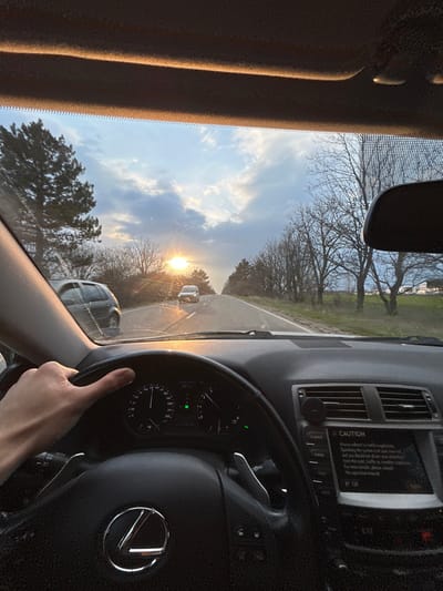 A first-person perspective image captures the interior of a Lexus vehicle and a rural road scene at sunset, ostensibly in Grigorevo, Bulgaria. A driver's left hand is positioned on the black leather steering wheel. The vehicle's instrument cluster indicates operation, and the central infotainment screen displays a "CAUTION" message related to traffic regulations. Directly ahead, the low setting sun causes significant glare and lens flare across the windshield, which exhibits multiple small streaks. The two-lane asphalt road stretches forward, marked with a solid white shoulder line. An oncoming white vehicle is visible in the distant lane. To the immediate left, a dark-colored SUV is partially visible, in the process of passing or having just passed the viewer's vehicle. The roadside environment features rows of bare deciduous trees and scattered evergreens under a partly cloudy sky, characteristic of a late winter or early spring evening.