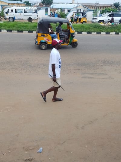 In Oja Oba, Oko Erin, Nigeria, a man walks on a dirt road, wearing a white t-shirt, beige shorts, and black and white sandals. He is holding a black stick in his right hand and has a red and white baseball cap on his head. Behind him, a yellow and black auto-rickshaw is on the road, with two people inside. To the left of the auto-rickshaw, a white van with blue stripes is parked. Further in the background, a silver SUV is parked on the side of the road, and a person is sitting near it. Buildings and trees are visible in the far background. The ground is covered in dirt and small debris.