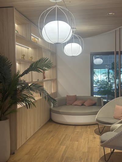 The image presents an interior space, likely a waiting or seating area within a building in Marcq-en-Barœul, France. The perspective is from a lower angle, capturing a partial view of the room's features.
On the left, there is a light-wood paneled wall with built-in shelving, some of which are illuminated. The shelves hold decorative items, including small figurines and what appears to be a perfume bottle.
Above the shelves, a modern, multi-bulb lighting fixture with spherical white shades hangs suspended. This complements the contemporary design of the space.
Moving towards the center, a potted plant with large, green fronds stands, partially obscuring the view of the shelving unit.
The upper right quadrant of the image showcases seating arrangements. A curved sofa with pale upholstery and pink cushions is visible, accompanied by two chairs made of wicker or a similar woven material.
A large window is present at the top, through which one can see exterior elements, including palm trees and architectural structures. The window suggests a tropical or subtropical theme, possibly reflected in the interior decor.
The floor is made of light-colored wood, adding warmth to the overall ambiance.