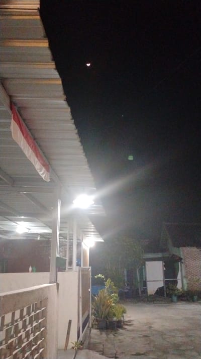 The image depicts an outdoor scene at night, likely in the vicinity of Banjarsari, Indonesia, given the red and white Indonesian flag hanging from a structure. In the dark sky, a crescent moon is visible. The foreground is dominated by a building structure with a corrugated metal roof, supported by a framework of metal poles. Bright lights illuminate the area under the roof, casting a glare. Part of a concrete wall with a decorative lattice pattern is visible on the left. Further into the scene, plants in pots are arranged on the ground, and a brick building is partially visible in the background. The ground appears to be paved. There are no people clearly visible in the image. The overall atmosphere suggests a residential or commercial outdoor area illuminated at night.