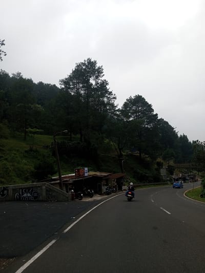 The image depicts a road winding through a hilly, forested landscape. A motorcyclist and a blue car are traveling along the road. A cluster of small shops or stalls sits adjacent to the road. Above, a hillside is covered with trees and green vegetation. The sky is overcast and gray. Graffiti is visible on a concrete structure beside the road. The setting appears to be in a rural or semi-rural area. The location given is the city of Mae Chan District, Thailand, but there is no specific information in the image to prove this location.