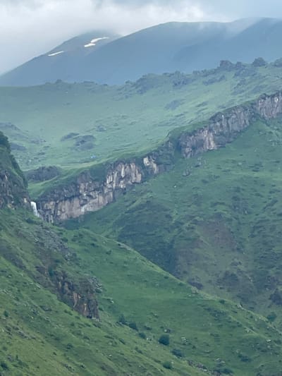 The image shows a panoramic view of a mountainous landscape in the vicinity of Arsha, Georgia. The scene is dominated by lush green hills and rugged rock formations, with a waterfall cascading down a cliff face. In the distance, misty mountain peaks rise against a cloudy sky, with patches of snow visible on one of the higher summits. The overall impression is one of natural beauty and rugged wilderness.