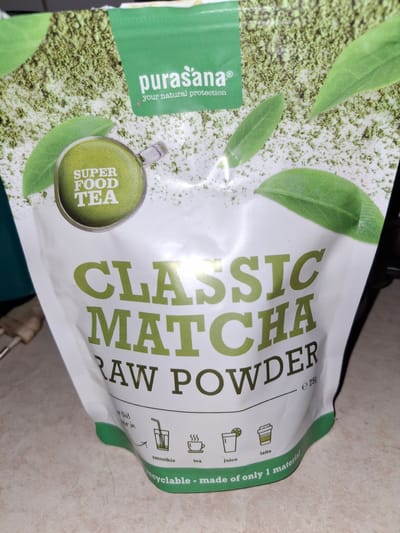 A close-up, eye-level, slightly angled shot shows a bag of Purasana Classic Matcha Raw Powder. The bag is predominantly white with green accents and text. The top half of the bag features a green band with the Purasana logo and "your natural protection" in white text. Behind this band, there's a texture resembling matcha powder interspersed with green leaves. A circular green graphic in the upper left reads "SUPER FOOD TEA." Prominently displayed across the middle of the bag in large, bold green lettering is "CLASSIC MATCHA RAW POWDER." Below this, the text "e 75g" is visible. The bottom portion of the bag shows small icons representing different ways to use the matcha: a smoothie, tea, juice, and latte. Text beneath these icons reads "recyclable - made of only 1 material." The bag is placed on a light-colored, textured surface that appears to be a countertop. The background is dark and out of focus. The image does not contain any specific elements indicating a location in Åsgårdstrand, Norway.