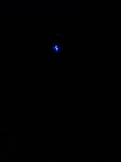 A dark, almost entirely black image is punctuated by a single, small source of blue light. It appears to be a tiny illuminated object, possibly a small LED or reflective element, positioned towards the upper-center area of the frame. There's a faint hazy line that is around the blue light. The rest of the composition is dark and indistinct, lacking detail.