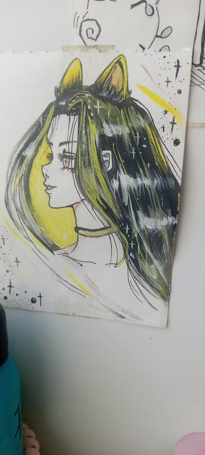 This is a close-up, medium-angle shot of an anime-style drawing, primarily featuring the profile of a young woman with cat ears. Her long, dark hair is streaked with yellow and white highlights, giving it a dynamic, flowing appearance. She has large, expressive eyes with reddish-brown irises, and her gaze is directed to the left. A thin, golden choker adorns her neck. The background of the drawing is a vibrant yellow circle, suggesting a halo or radiant aura. Scattered across the drawing are small white and black crosses and dots, resembling stars or celestial elements. The drawing is done on a piece of paper that appears to be taped to another surface, as indicated by a strip of beige tape visible at the top. The paper itself shows some signs of age and wear, with faint smudges and a slightly textured surface. The image is taken against a plain white background, with a sliver of a turquoise bottle and a hint of pink material visible in the bottom left corner. The overall mood of the drawing is somewhat mystical and ethereal, with a hint of playful fantasy due to the cat ears.