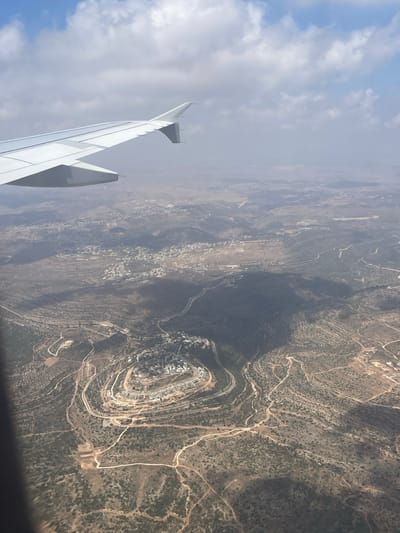 An aerial view from an airplane shows a landscape with scattered settlements and winding roads through hilly terrain. A prominent settlement, appearing as a cluster of white buildings arranged in a circular pattern on a hilltop, is visible in the center-right of the image. The surrounding hills are covered with sparse vegetation and crisscrossed by dirt roads. The shadow of the airplane wing is cast over a portion of the landscape. The sky above is partly cloudy. The image is taken from a high altitude, providing a broad perspective of the area.