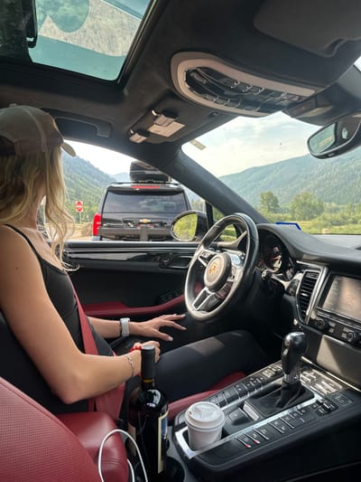 The image depicts the interior of a dark-colored Porsche Macan from the front passenger perspective, looking forward on a clear day in Aspen, United States. A blonde individual, wearing a light-colored baseball cap, black tank top, and black leggings, is seated in the driver's seat, secured by a prominent red seatbelt. A white smartwatch is visible on their left wrist, and a red bracelet on their right wrist. A dark red wine bottle stands upright in a cup holder on the red-accented center console, adjacent to a white disposable coffee cup with a lid in another cup holder. A white charging cable is visible near the driver's seat. Through the windshield, a black Chevrolet SUV, equipped with a roof-mounted cargo box and a rear hitch rack, is directly ahead, appearing to be stopped or moving slowly. A red 'Do Not Enter' sign is visible on the roadside to the left of the lead SUV. The background features steep, green, tree-covered mountains under a bright, hazy sky.