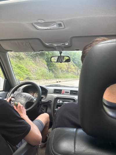 The image captures the interior of a Honda vehicle, specifically from the perspective of a rear passenger, looking towards the driver and the road ahead in Нареченски бани, Bulgaria.
The driver, a male wearing a black t-shirt and grey shorts, has both hands on the black, four-spoke steering wheel with a Honda emblem. His left hand grips a small, olive-green object. Car keys are in the ignition. The grey dashboard features a visible instrument cluster and a Pioneer car stereo in the center console. A red hazard light button is present above the stereo. The driver's sun visor is lowered, displaying a warning label.
The back of a front passenger's head, with brown hair, is visible on the right, partially obscured by a black headrest. This passenger is wearing dark clothing.
Beyond the windshield, an asphalt road winds through a natural environment. A concrete barrier lines the right side of the road, separating it from a steep hillside covered in dense green vegetation, including trees and bushes, and exposed rock formations. The scene is illuminated by bright daylight, suggesting clear weather conditions.