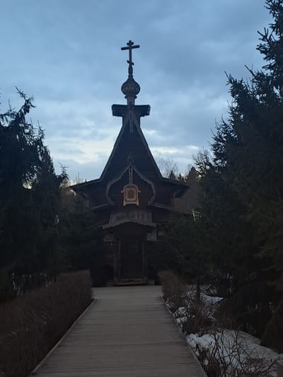 The image depicts the Church of the Intercession of the Holy Virgin, located in Vzglyadnevo, Russia. The church is a wooden structure with a distinctive onion dome topped with a cross. The building is framed by evergreen trees. A wooden boardwalk leads to the church entrance. The sky is overcast.