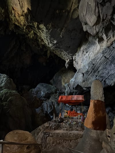 The image depicts the interior of a cave in Hong Ngern Village, Laos. A reclining Buddha statue, draped in orange fabric, is positioned on a raised platform adorned with offerings. The cave walls and ceiling feature natural rock formations, including stalactites and stalagmites, with areas illuminated by artificial light. The scene suggests a place of worship within a natural cavern.