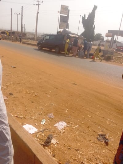 The image displays a roadside scene in Lapai, Nigeria, under daylight conditions with a hazy sky. A dark blue minivan, overloaded with various items secured to its roof and rear, is parked on the right side of a paved road. Six individuals are visible in the vicinity of the minivan. One person in a yellow outfit and another in a light-colored top are positioned at the rear of the vehicle, actively appearing to load or unload items. Three other individuals in various light-colored shirts stand nearby. Further to the right, a sixth person, also in a yellow outfit, walks away from the minivan while carrying a bag. A seventh individual is seen walking on the far left side of the paved road. The immediate foreground consists of a reddish-brown, unpaved dirt shoulder, extensively littered with plastic bags, paper, and miscellaneous debris. A concrete curb is present along the bottom left edge of the frame. In the background, a Petroburg/TOTAL petrol station is identifiable by its sign and canopy structure, located behind the minivan. Multiple utility poles carrying power lines extend along the road, with various residential buildings visible on elevated ground in the distance.
