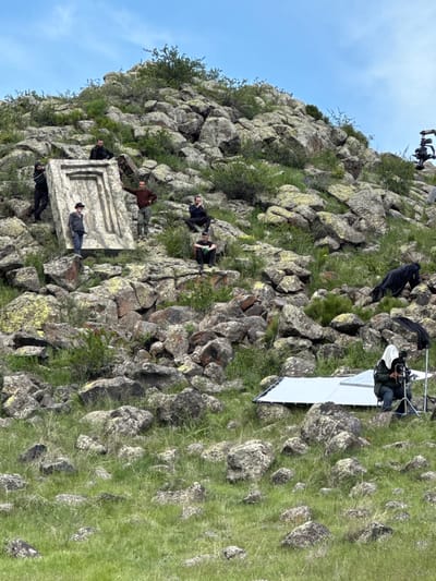 The image depicts a film set on a rocky hillside, likely in a rural area. People are present, some appearing to be crew members operating cameras and lighting equipment, while others seem to be actors positioned near a rectangular stone structure with a raised border. The structure is sitting on the hillside. The environment consists of rocks, green vegetation, and a clear blue sky. The location is Tlik, Armenia. The action appears to be a film shoot.