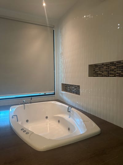 This image depicts a clean, unoccupied modern bathroom located in Ban Ko Li Pe, Thailand. The central subject is a large, white, rectangular jacuzzi bathtub with rounded corners, positioned on a dark brown floor, possibly laminate or large tiles. The bathtub features multiple visible jets, chrome faucets with a shower wand on its left edge, and two chrome grab handles on its rim. Three circular white control buttons are present on the tub's front-facing edge.
The walls are covered with white vertically-textured tiles. Integrated into the tiled wall on the right are two recessed niches, each backed with a mosaic pattern of small rectangular tiles in shades of brown, black, and white. To the left, a large window is covered by a light-colored (beige/off-white) roller blind, obscuring the view outside. The white ceiling contains a single illuminated recessed spotlight in the upper left corner, casting light into the room. No individuals or personal items are present in the scene.