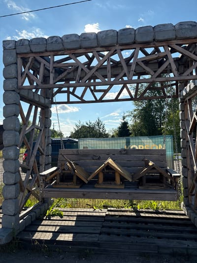 The image shows a rustic wooden structure, possibly an outdoor seating area or play structure, set in an outdoor environment. The structure is made of rough-hewn wood beams and planks, with a stone-like decorative element along the base of some sections. Chains are used to suspend parts of the structure. In the background, there are trees, a blue sky with white clouds, and what appears to be a glass-like barrier with yellow text on it. The lighting suggests it is daytime, with sunlight casting shadows. The overall impression is of an informal, possibly handmade, outdoor installation. Visible text appears on a panel in the background, possibly indicating a name or location, with letters like "OP" and "ON" discernible. The scene appears to be in a rural or semi-rural setting, given the natural elements and the rustic construction. There are no people visible in the image. The time of day appears to be late afternoon, given the angle of the sun and the shadows. The weather is clear with some scattered clouds.