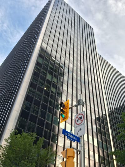 This is an upward-angle shot of a modern, tall building with a reflective facade. The building's exterior is composed of many horizontal slats, creating a ribbed effect. The facade reflects the sky and clouds, which are partially visible at the top right of the image, suggesting it's daytime. A large green tree is in the foreground on the left, partially obscuring the building's lower section.
Below the tree and to the right, a traffic light is visible. The traffic light shows a green light illuminated. A blue street sign is attached to the traffic light pole, reading "rue Queen St." with an arrow pointing left and down. Next to the street sign are two regulatory signs. The first sign shows a black arrow curving left. The second sign is a circular prohibition sign with a red border and a diagonal red bar across an image of a cigarette, indicating "no smoking."
The scene appears to be a city street in Ottawa, Canada, given the street name and the style of the building and traffic signals. The lighting suggests it is daytime, possibly late afternoon or early evening, with a partly cloudy sky. The overall mood is neutral, focusing on the architectural elements and urban infrastructure.