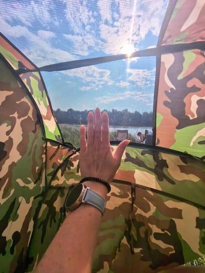 A person's hand, with bright yellow fingernails, rests against a fine mesh screen, obscuring a view of a sunny day. The mesh is part of a camouflage-patterned tent, with patches of green, brown, and tan visible. The hand is adorned with a black coiled bracelet and a smartwatch with a light blue strap and a silver casing. Beyond the mesh, a body of water is visible, with trees lining the distant shore. Two figures, one partially visible, are standing near the water. The sky above is a bright blue, dotted with white clouds, and the sun shines brightly through the mesh, creating lens flare. The image is taken from inside the tent, looking outwards. The location is identified as Kuzminovka, Russia.