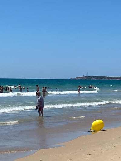 This image depicts a beach scene in Conil de la Frontera, Spain. The foreground is dominated by a sandy beach with a yellow buoy partially submerged at the water's edge. In the middle ground, several people are visible wading and swimming in the ocean, with waves breaking on the shore. A man wearing a light-colored shirt and red shorts stands in the shallow water, holding a dark object. The background shows more people in the sea and a distant coastline with structures, possibly lighthouses, against a clear blue sky. The water is a vibrant turquoise, suggesting a sunny day.