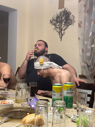 The image shows a man sitting at a table with food and drinks. He is wearing a black shirt, and his head is wrapped in white napkins or cloth, with a fork sticking out of the top. He is holding a fork with a few ring-shaped snacks on it to his mouth. There are several glasses on the table, some filled with liquid, as well as beer cans and plates with food. The background includes a curtain and a decorative piece on the wall resembling a branch. Given the "пиринско" label on the beer cans, and the prompt mentioning Galiche, Bulgaria, it is reasonable to assume this scene takes place there.