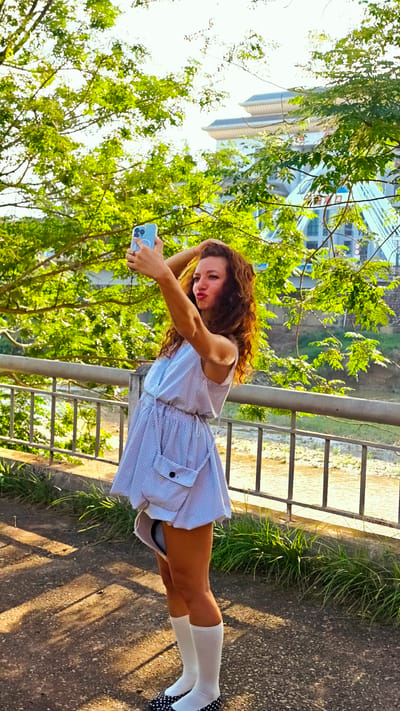 A young Caucasian woman with long, wavy auburn hair stands on a paved walkway next to a metal railing in Lao Cai Ward, Vietnam. She is dressed in a short, blue and white checkered dress with a large pocket, white knee-high socks, and black and white polka-dot shoes. The woman is holding a light blue smartphone up at arm's length, presumably taking a selfie, and has a pouting expression on her face. A bridge and a building with a tiered roof are visible through the green foliage in the background. A river is seen through the metal railing. The photograph is taken in bright daylight.