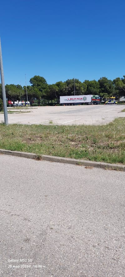 The image depicts a parking area located near the city of l'Aldea, Spain. A series of commercial vehicles, including trucks with trailers, are parked along a roadside bordered by trees. A large sign with the text "A. BELZUNCES" is prominently displayed on one of the trailers. The ground consists of asphalt and a grassy area, with a clear blue sky visible above the trees. The image was taken on May 26, 2026, at 16:06.