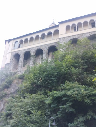 The image shows a building with arched windows and a cloister-like structure, set against a backdrop of lush green trees and a rocky cliff. The building appears to be on a hillside. The sky is overcast and bright white, suggesting it is daytime. In the foreground, a lamppost is visible. The architecture and the surrounding vegetation suggest a location with a temperate climate and possibly historical significance. The building's design, with its series of arches and its positioning on a steep incline, could indicate it is a monastery or a similar institution, potentially with a view. The overall impression is one of a serene, somewhat enclosed, and possibly remote setting.