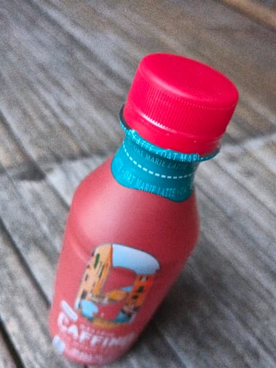 A reddish-pink plastic bottle with a bright red ribbed screw cap is positioned diagonally across a wooden surface. A teal-colored neckband below the cap displays repeating white text, partially legible as "OAT MARIE LATTE," with a dashed white line beneath. A portion of a label on the bottle's body features a graphic illustration of multi-story buildings, possibly with a canal, and partially visible white text "CAFFINO." The bottle rests on a greyish-brown, weathered wooden surface composed of planks. No people are present. No specific location in Kyauk Hsoke, Myanmar, can be determined from the visual evidence provided.