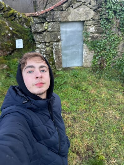 The image captures a young adult male taking a selfie outdoors in Ribeira de Pena, Portugal. The individual, positioned in the foreground, has light brown hair, blue eyes, and light facial stubble, looking directly at the camera with a neutral expression. He wears a black hooded sweatshirt beneath a dark blue quilted puffer jacket. The background features an old stone wall, constructed from irregular grey masonry, partially covered with green moss and ivy on the right side. Within this wall, a rectangular opening is framed by larger stones and sealed by a plain, light grey metallic door. A small white rectangular sign is visible on the moss-covered section of the wall to the left of the door. The ground between the individual and the wall is covered in sparse green and brown grass. Bare tree branches are discernible above the wall in the upper background, indicating an outdoor setting under what appears to be an overcast sky.