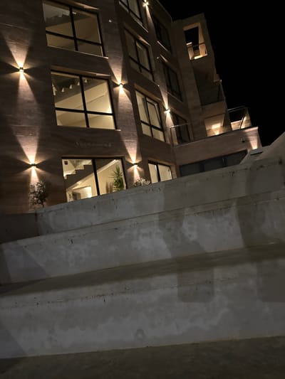 The image shows a modern building exterior at night, likely in Tyulenovo, Bulgaria. The building has a textured facade, large windows, and is illuminated by exterior wall lights. In the foreground, there are concrete steps or a tiered walkway. The steps are weathered and appear to be made of a rough-textured material. The building has multiple stories, with balconies or terraces visible on the upper levels. The overall impression is one of contemporary architecture set against a dark night sky.